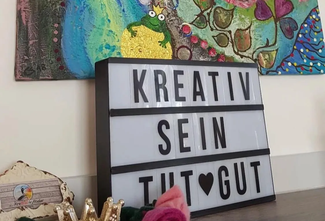Leuchtschild mit den Worten „Kreativ sein tut gut“ vor bunten Bildern